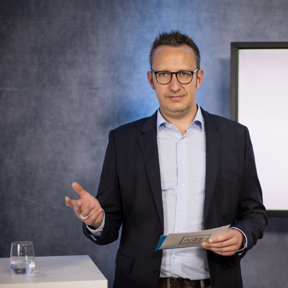 AV22 film – Philipp Klietz steht am Moderationstresen und hält eine Moderationskarte in der Hand