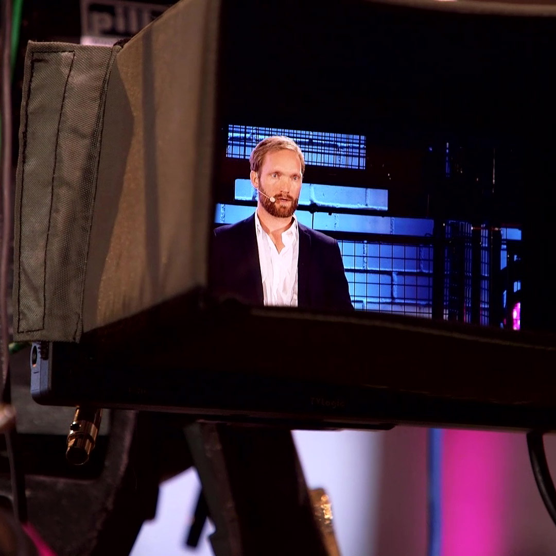 AV22 film – Ein Redner mit Mikrofon ist durch das Display einer Kamera zu sehen